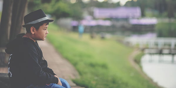 Boy in Black Jacket and Black Fedora Hat Sitting Near Body of Water thumbnail