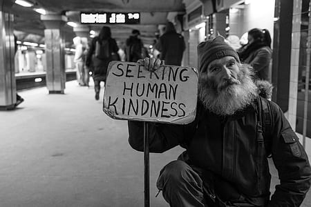 grayscale photo of man kneeling on ground holding seeking human kindness signage at subway station thumbnail
