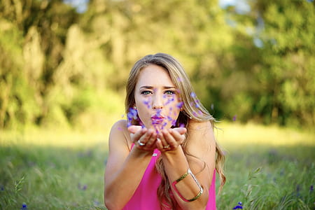 woman blowing purple flower petals near green trees thumbnail