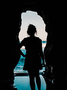 silhouette of person standing inside cave near seashore during daytime thumbnail