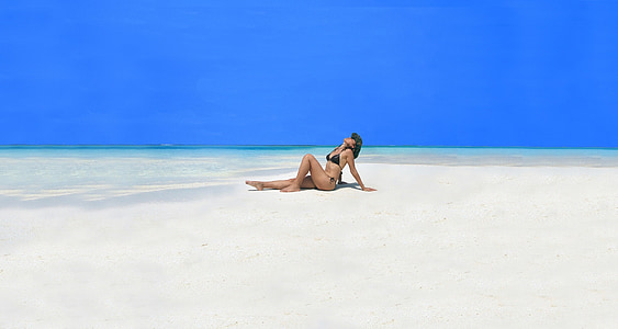 woman wearing black bikini sitting on sand near seashore with hands behind back and looking upwards thumbnail