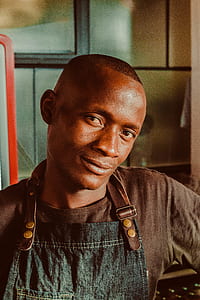 man wearing brown crew-neck shirt and blue apron slightly smiling while facing camera direction closeup photo thumbnail