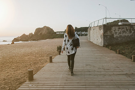 woman in white and black star print long-sleeved shirt and black denim jeans walking on brown wooden pathway near body seashore thumbnail