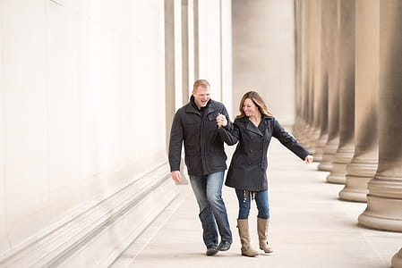 Man And Woman Wearing Black Coat And Jacket Walking Near Tall Pillars thumbnail