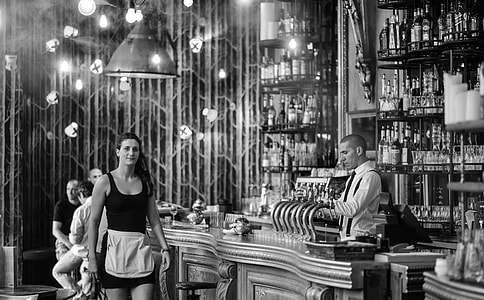 grayscale photography of man wearing white dress shirt while standing near black metal bottle rack filled with bottles thumbnail