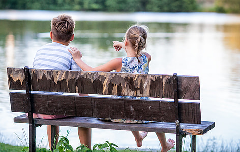 girl and boy sitting on brown wooden bench near body of water at daytime thumbnail