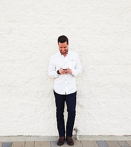 man wearing white dress shirt and black dress pants holding smartphone while leaning to white wall thumbnail
