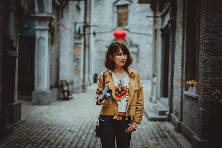 woman standing beside brown concrete building during daytime while carrying gray DSLR camera thumbnail