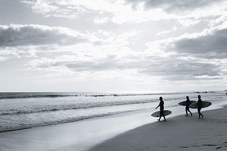 grayscale photo of three people holding surfboards walking on seashore thumbnail