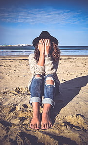 woman in gray sweater and blue denim distressed pants covering face with hands sitting on sand near body of water thumbnail