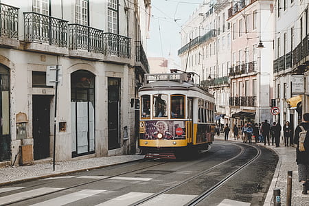 yellow and white tram in the middle of road surrounded by people walking around thumbnail