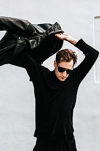 man wearing black leather jacket standing near white painted wall thumbnail