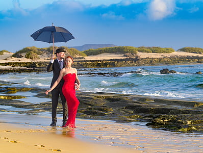 man in black suit holding black umbrella with woman in red dress on beach thumbnail
