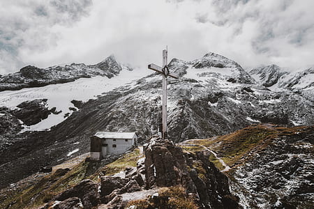 photo of cross on rocky hill near white concrete building and snow-covered mountain thumbnail