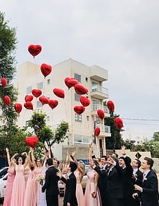 Women Wearing Pink Dresses and Men Wearing Black Suit Jacket and Pants Raising Hands With Red Heart Balloons thumbnail