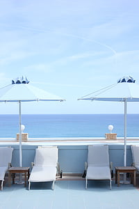 four white outdoor loungers under shade of two blue parasols overlooking sea under clear blue sky during daytime thumbnail