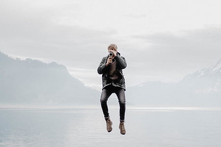 man jumping while holding camera with calm body of water background thumbnail