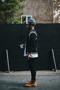 man wearing black and white jacket and black pants standing near black wall at daytime thumbnail