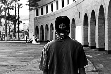 grayscale photography of person wearing cap standing beside structure thumbnail