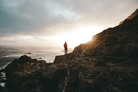 person standing on mountain cliff facing body of water under cloudy sky thumbnail