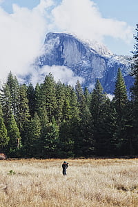 man taking photo while standing on withered grass field under snow capped mountain thumbnail