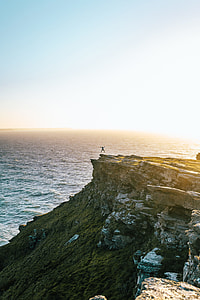 person jumping on cliff near body of water during golden hour thumbnail