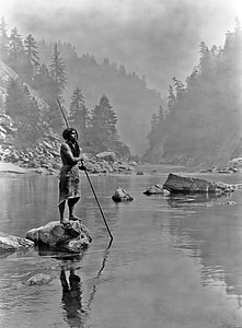 topless man standing on rock while holding rod in mist river thumbnail