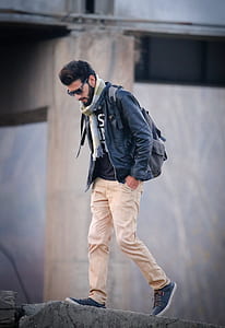 man in blue jacket and brown pants with backpack standing on concrete near building thumbnail