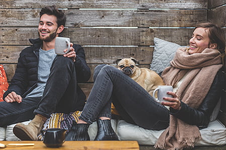 man wearing black zip jacket sitting beside woman with brown scarf and fawn pug on woman's lap thumbnail