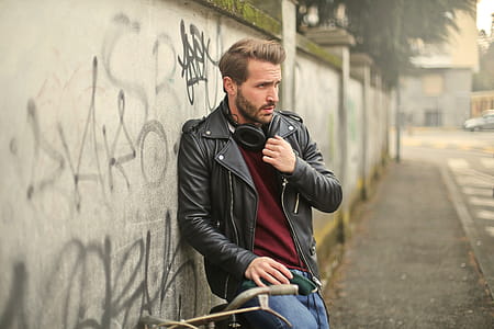 A young caucasian man wearing a leather jacket and jeans sitting against a wall on a bicycle thumbnail