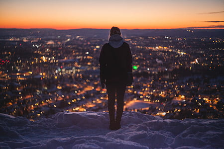 woman wearing hoodie standing near the edge of snow covered cliff facing the city during night thumbnail