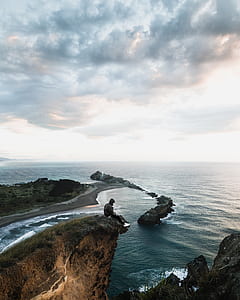 man sitting on cliff in front of body of water thumbnail