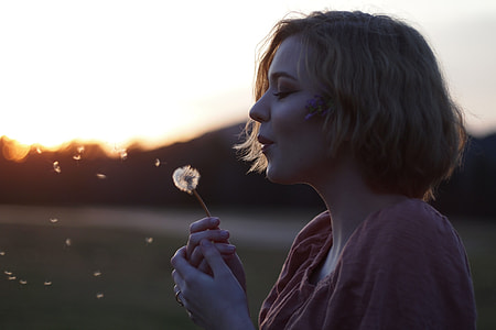 woman blowing white dandelion flower on open field under white and yellow skies thumbnail