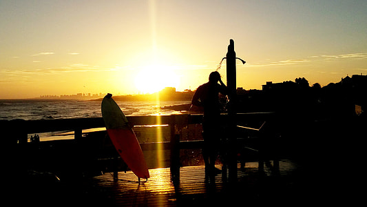 silhouette of man standing on the shower near surfboard and sea shore during sunset thumbnail