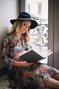 Pregnant Woman Wearing Green, Red, and White Floral Dress Reading a Book Near Window thumbnail
