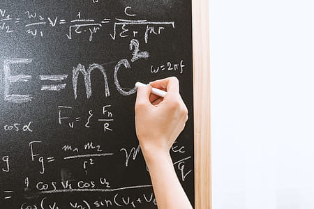 Female hand holding white chalk in front of a blackboard with Einstein formula. Education and school concept. thumbnail