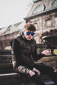 man in black sweater and brown camouflage hooded vest smokes a cigarette and sits on brown stairs during daytime thumbnail