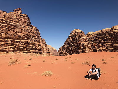 Man Wearing White Shirt and Black Pants Sitting on Soil Behind Rock Formation Mountain thumbnail