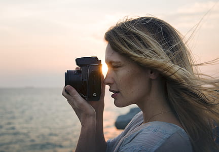 portrait photography of woman holding black DSLR camera near body of water during daytime thumbnail