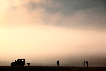 silhouette photo of man taking photo through camera with tripod stand of man standing and black vehicle under cloudy sky thumbnail