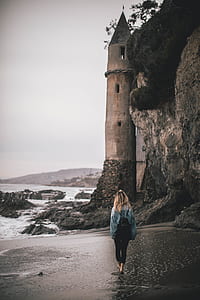woman walking in between seashore and hill towards gray concrete tower thumbnail
