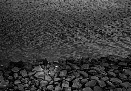 grayscale photography of man standing on rocks near body of water thumbnail