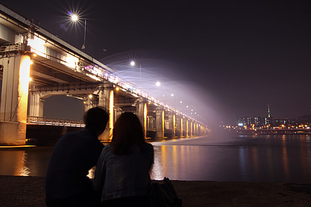 man and woman near lighted bridge and ocean during nighttime thumbnail