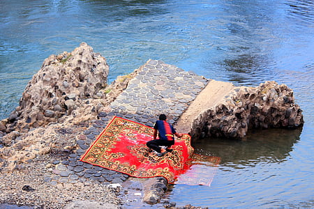 man washing carpet on rock beside body of water thumbnail