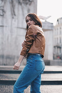 woman wearing brown leather jacket, blue jeans standing near body of water in closeup photography thumbnail