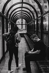 grayscale photography of woman walking in front man sitting on chair at hallway thumbnail