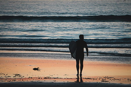 man walking on seashore while holding surfboard thumbnail