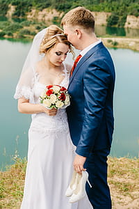 Groom Holding a Pair of White Leather Heeled Shoes Standing in Front of the Bride in White Wedding Gown thumbnail