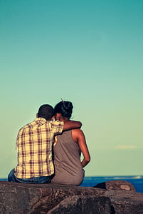 man wearing beige and gray plaid shirt hugging woman in gray tank top sitting on gray concrete ledge thumbnail