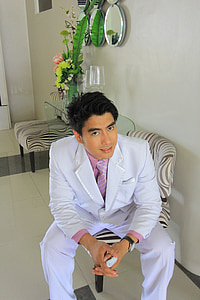 man in white suit sitting on gray chair posing for photo thumbnail